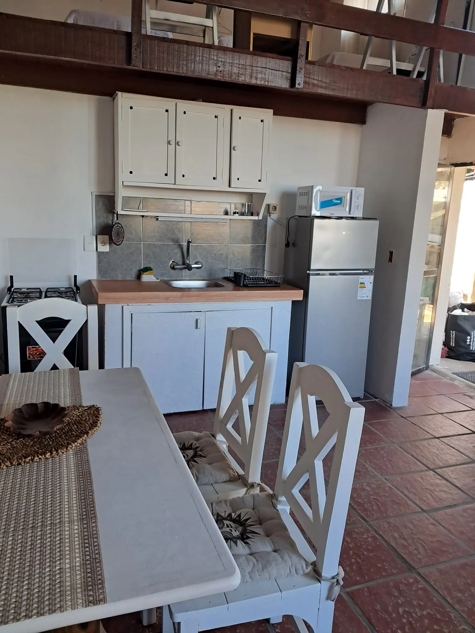 Cocina con hornallas a gas, heladera, microondas y mesa con sillas blancas, bajo un entrepiso de madera