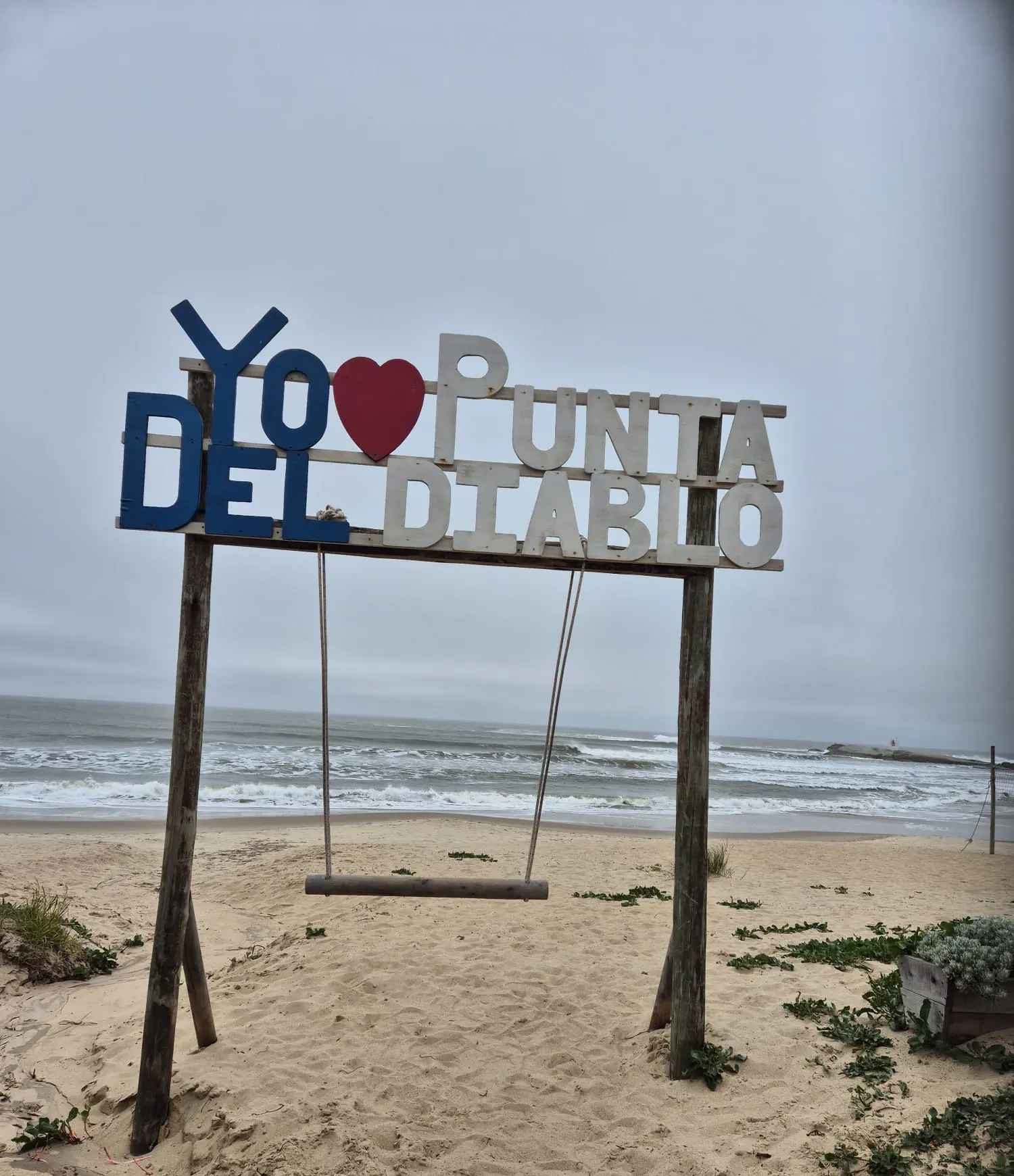 Cartel «Yo amo Punta del Diablo» con una hamaca de madera sobre la arena, frente al mar