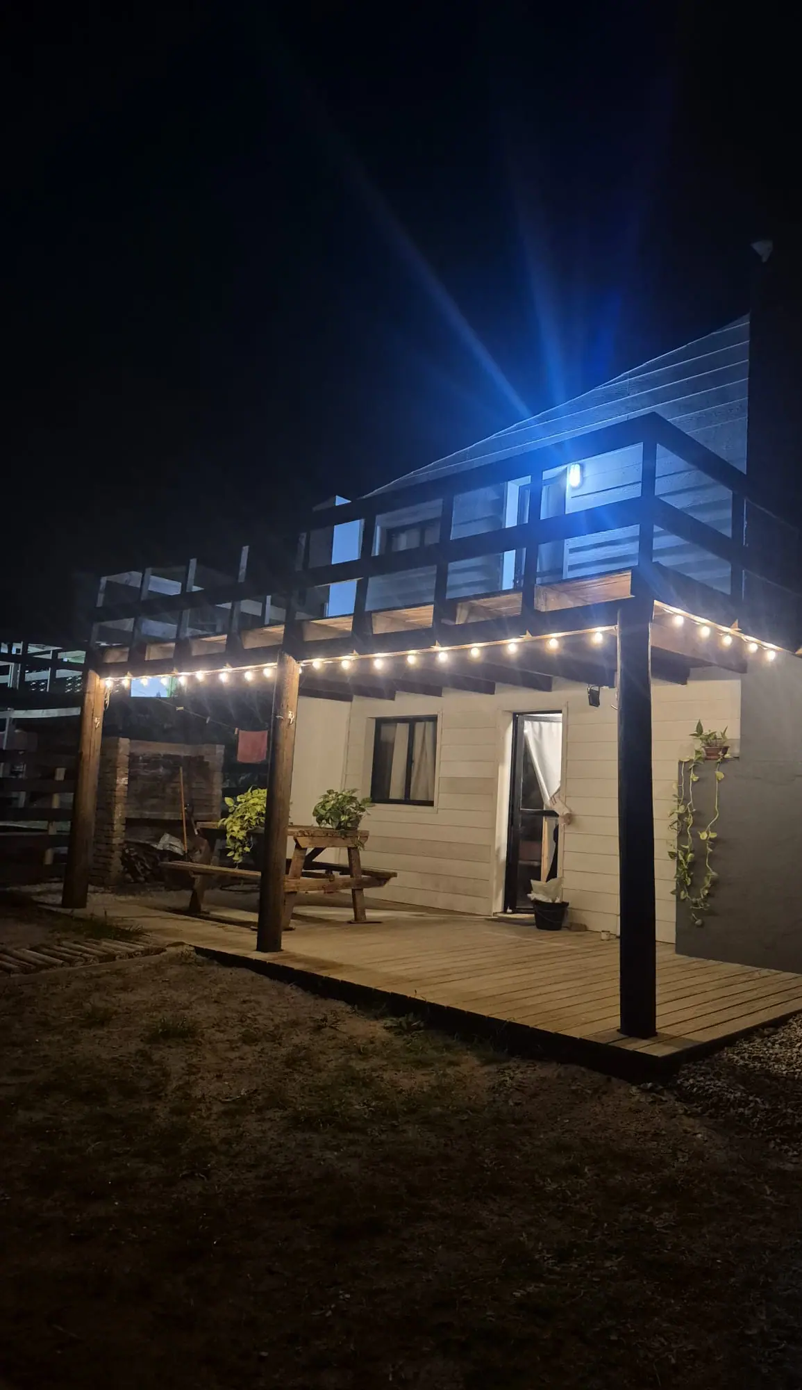 La casa de noche, con una guirnalda de luces sobre el deck de madera y una mesa de picnic